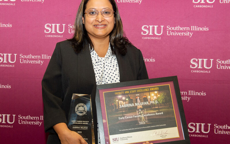 A woman with dark hair and glasses, smiling, and holding an award and framed certificate. Behind her, the wall has the SIU logo.