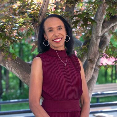 A woman wearing a red, sleeveless dress, is standing in front of a tree.