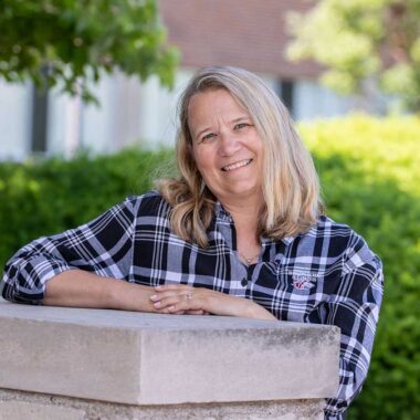 A woman is standing behind a grey brick wall. It comes up to her chest. She has her arms resting on the ledge, and she is smiling. She has blond hair and is wearing a black and white shirt.