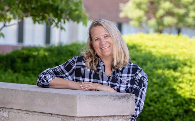 A woman is standing behind a grey brick wall. It comes up to her chest. She has her arms resting on the ledge, and she is smiling. She has blond hair and is wearing a black and white shirt.