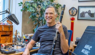 A man is seated, and smiling, while holding a clarinet. He is wearing a blue t-shirt and jeans. There is a potted plant in the background.