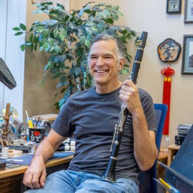 A man is seated, and smiling, while holding a clarinet. He is wearing a blue t-shirt and jeans. There is a potted plant in the background.