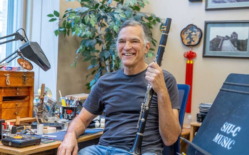 A man is seated, and smiling, while holding a clarinet. He is wearing a blue t-shirt and jeans. There is a potted plant in the background.