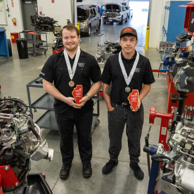 Two students stand in an auto shop, holding awards.