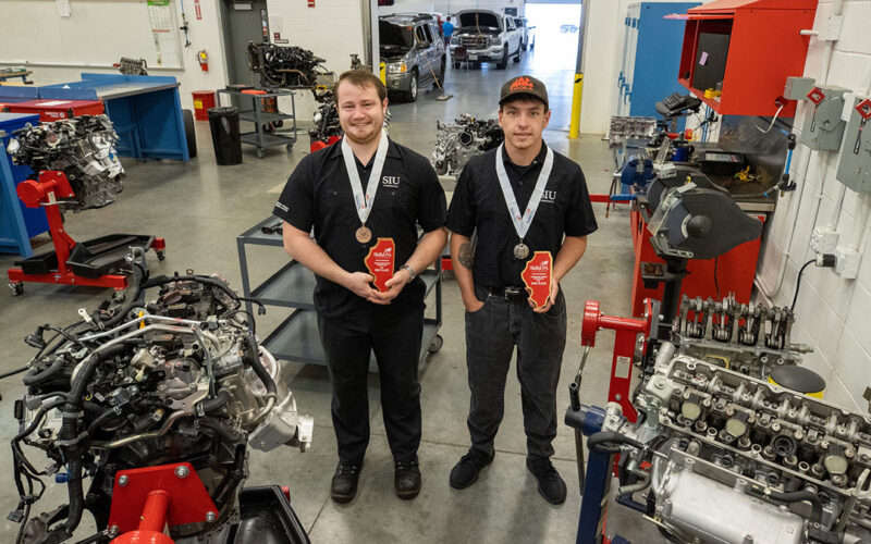 Two students stand in an auto shop, holding awards.